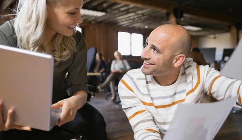 Blonde-haired woman rests a laptop in her hands as she chats to a bald man dressed in a stripy white jumper.